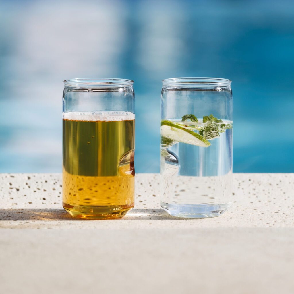 Two Govino shatterproof beer can glasses filled with a beer and a cocktail in front of a pool outdoors